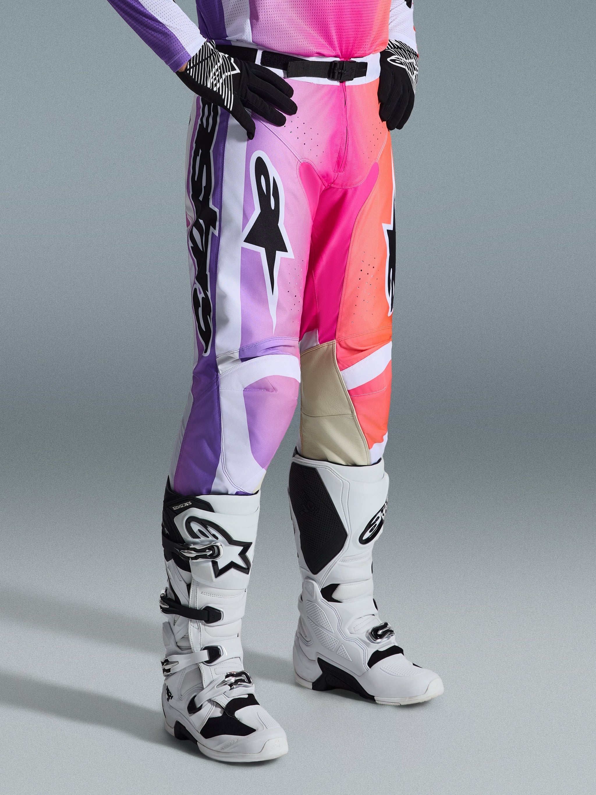 Pantalon Alpinestars Racer Air Portl 2026, pantalon de motocross, graphisme multicolore avec dégradés de violet, rose et orange, porté avec des bottes de motocross blanches et noires et des gants noirs, doté d'une coupe de course et de renforts intérieurs aux genoux en cuir beige.