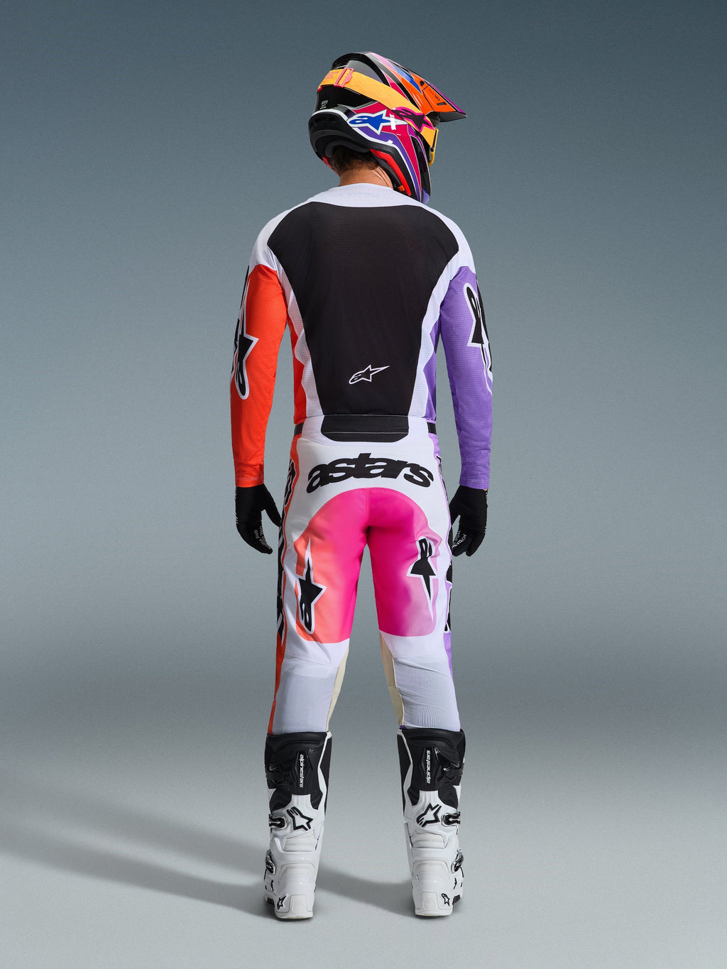 Vue arrière d'un pilote de motocross portant le pantalon Alpinestars Racer Air Portl 2026 en version Multicolore, caractérisé par un tissu blanc avec des panneaux dégradés rose et orange et des logos noirs, associé à un maillot noir aux manches violettes et orange, des bottes de course blanches et un casque multicolore éclatant.