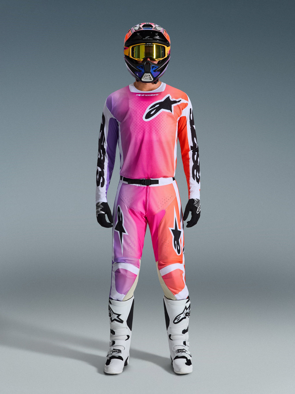 Tenue de motocross complète Alpinestars Racer Air Portl 2026, avec dégradé rose, violet et orange sur le pantalon et le maillot, casque assorti, lunettes jaunes, bottes blanches et gants noirs pour un look tout-terrain coordonné.