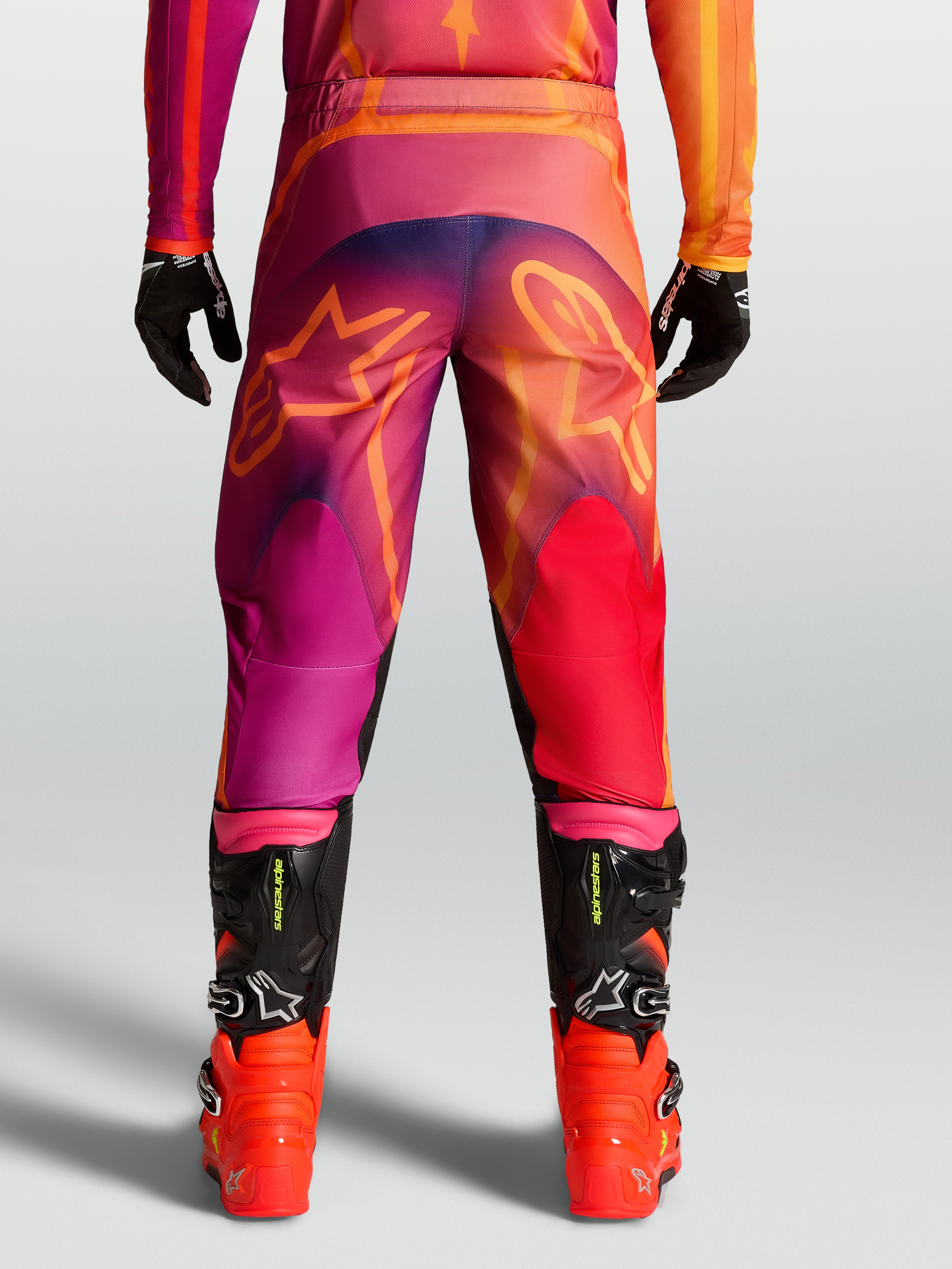Pantalon Fluid Kepler d'Alpinestars, pantalon de motocross pour homme en violet et orange avec motifs d'étoiles à l'arrière et bottes de racing noires.