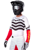 Pilote de motocross portant le maillot Alpinestars Limited Edition Techstar Deep, maillot blanc avec rayures ondulées noires et bas de manches rouges, assorti à un casque noir, des lunettes miroir, des gants blancs et noirs et un pantalon de course blanc, tenue de motocross haute performance.