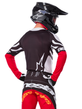 Pilote de motocross vu de dos portant le maillot Alpinestars Limited Edition Techstar Deep, noir, blanc et rouge vif, avec un pantalon de course assorti et un casque noir, coupe athlétique pour la performance tout-terrain