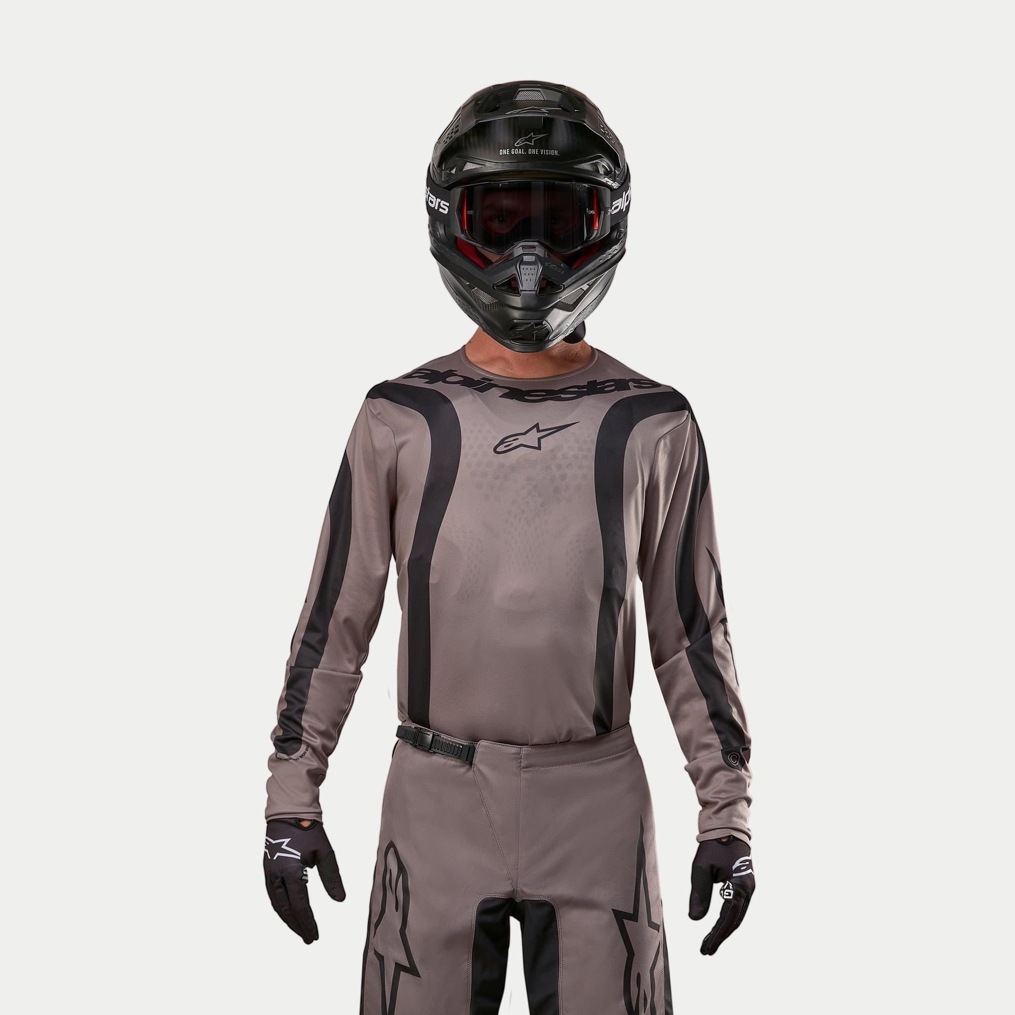 Motocross rider wearing the Alpinestars 2024 Fluid Lurv Jersey in Mud Black, an earthy grey-brown fabric with black graphic stripes and a star logo on the chest, paired with a black helmet, red goggles, and black gloves for a coordinated off-road racing look