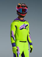 Pilote de motocross portant le maillot Alpinestars 2026 Supertech Vista, jaune vif avec des accents violets et noirs, pantalon et gants jaunes assortis, casque multicolore avec lunettes violettes, doté de perforations au laser et d'un grand logo Astar sur la poitrine, tenue de course coordonnée de niveau professionnel
