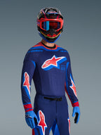 Pilote de motocross portant le maillot Supertech Vista 2026 en bleu marine, corail et cobalt, pantalon et gants assortis, casque multicolore avec lunettes rouges, équipement professionnel pour la course tout-terrain