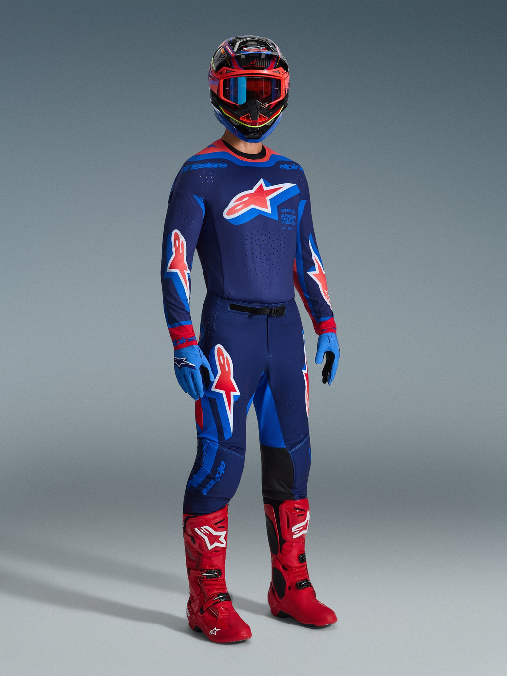 Pilote de motocross portant une tenue de course complète, maillot Supertech Vista 2026 bleu marine foncé avec accents corail vif et cobalt, pantalon de motocross assorti, casque multicolore avec lunettes rouges, bottes rouge vif, gants bleus, équipement coordonné pour la course tout-terrain
