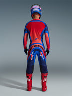 Pilote de motocross vu de dos portant le maillot Alpinestars Supertech Vista 2026 en Dark Navy, Hot Coral et Cobalt, tenue complète avec pantalon assorti, bottes de course rouges et casque bleu, équipement de course tout-terrain professionnel.