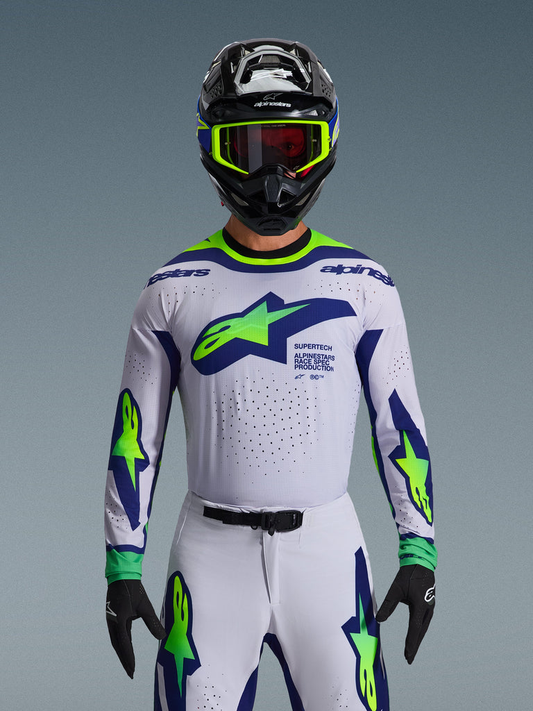 Maillot Alpinestars Supertech Vista 2026, maillot de motocross, gris clair, jaune fluo et bleu, porté par un pilote avec pantalon assorti et casque noir, avec un large logo sur la poitrine et des perforations au laser pour la ventilation, coupe athlétique de niveau professionnel