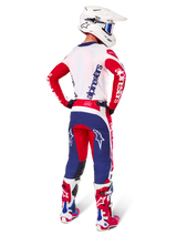Pilote de motocross vu de dos portant le maillot Alpinestars Limited Edition Techstar Fortitude, coloris rouge vif, blanc et bleu, pantalon technique assorti avec logos d'étoiles, casque blanc avec lunettes et bottes rouges et blanches, équipement complet coordonné pour la course tout-terrain