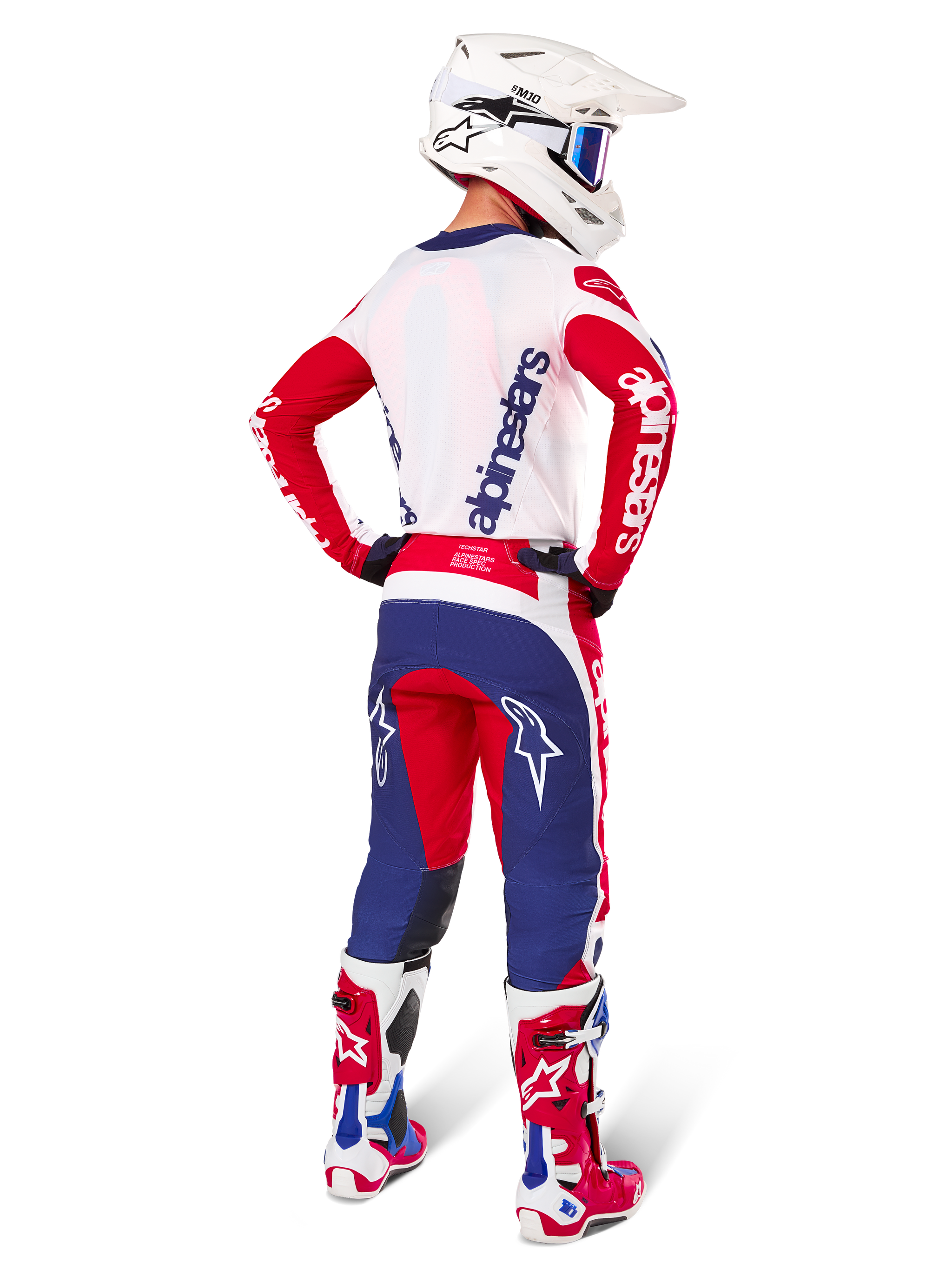 Pilote de motocross vu de dos portant le maillot Alpinestars Limited Edition Techstar Fortitude, coloris rouge vif, blanc et bleu, pantalon technique assorti avec logos d'étoiles, casque blanc avec lunettes et bottes rouges et blanches, équipement complet coordonné pour la course tout-terrain