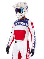 Pilote de motocross portant le maillot Alpinestars Limited Edition Techstar Fortitude, rouge vif, blanc et bleu, avec un casque blanc, des lunettes bleues et un pantalon assorti, équipement de course haute performance pour la compétition tout-terrain