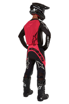 Pilote de motocross vu de derrière portant le maillot Alpinestars Supertech Ward 2024 en Black Red Berry, avec un panneau dorsal rouge vif et des manches noires, associé à un pantalon assorti, des bottes Tech 10 noires et un casque en fibre de carbone pour une tenue de course professionnelle.