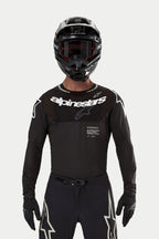 Pilote de motocross portant le maillot Alpinestars Supertech Ward 2024 noir, coupe de course athlétique avec logo Alpinestars blanc sur la poitrine, perforations découpées au laser et inscription 'Supertech Race Spec', associé à un casque et des lunettes noirs pour la course tout-terrain