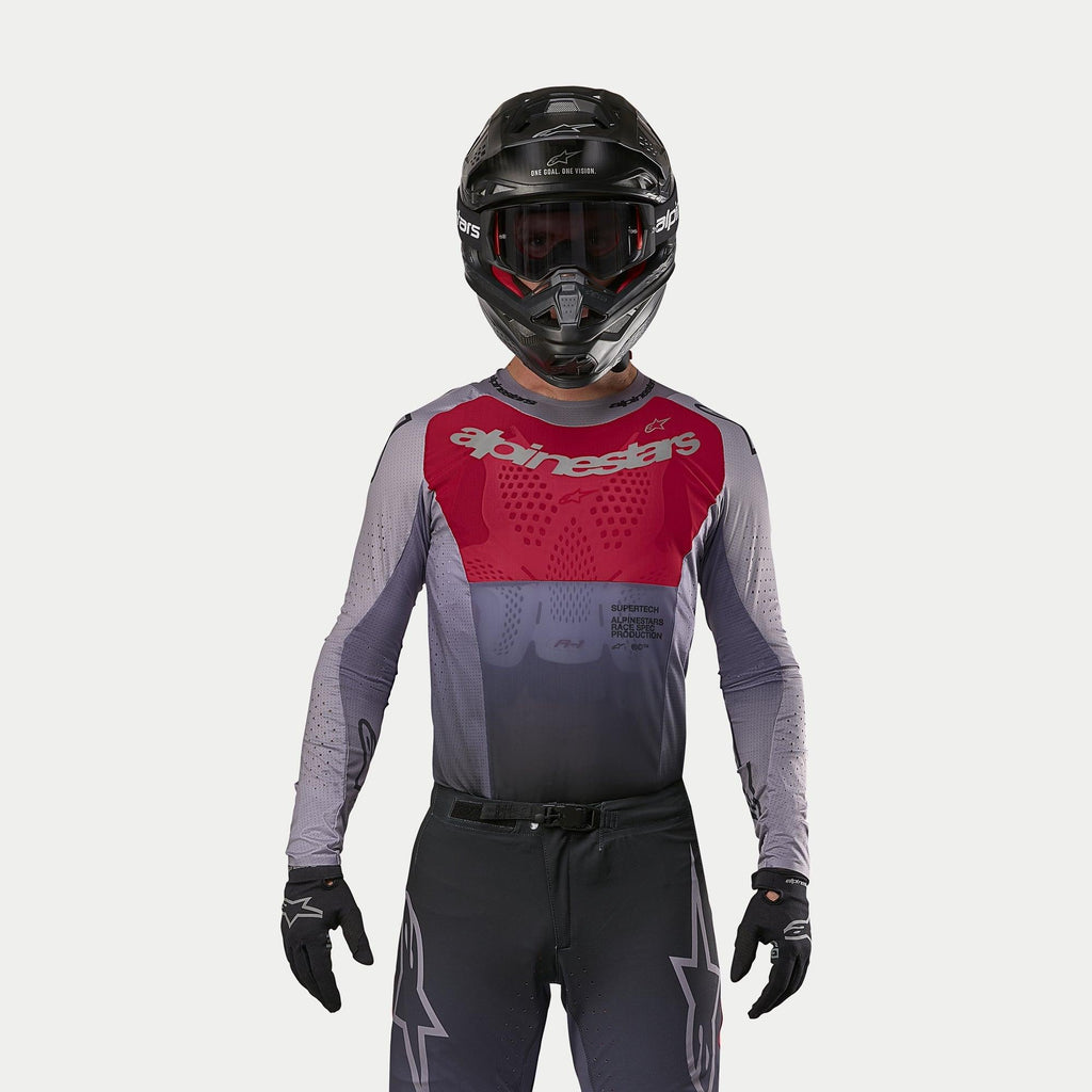 Pilote de motocross portant le maillot Alpinestars Supertech Dade 2024 en Iron Red Berry, doté d'un panneau rouge sur la poitrine et de manches grises avec logos argentés, porté avec un pantalon assorti, un casque noir et des gants noirs, équipement de course tout-terrain professionnel