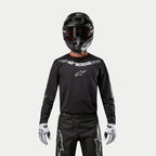 Maillot Alpinestars Fluid Graphite 2024, maillot de motocross noir et argent, porté par un pilote avec un casque noir et des lunettes rouges, logo Alpinestars argenté sur la poitrine, coordonné avec un pantalon noir et des gants blancs pour la performance tout-terrain