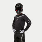 Pilote de motocross portant le maillot Alpinestars Fluid Graphite 2024 noir et argent, associé à un pantalon de motocross noir, un casque noir avec des lunettes teintées de rouge et des gants blancs et noirs, avec des logos argentés sur la poitrine et les manches.