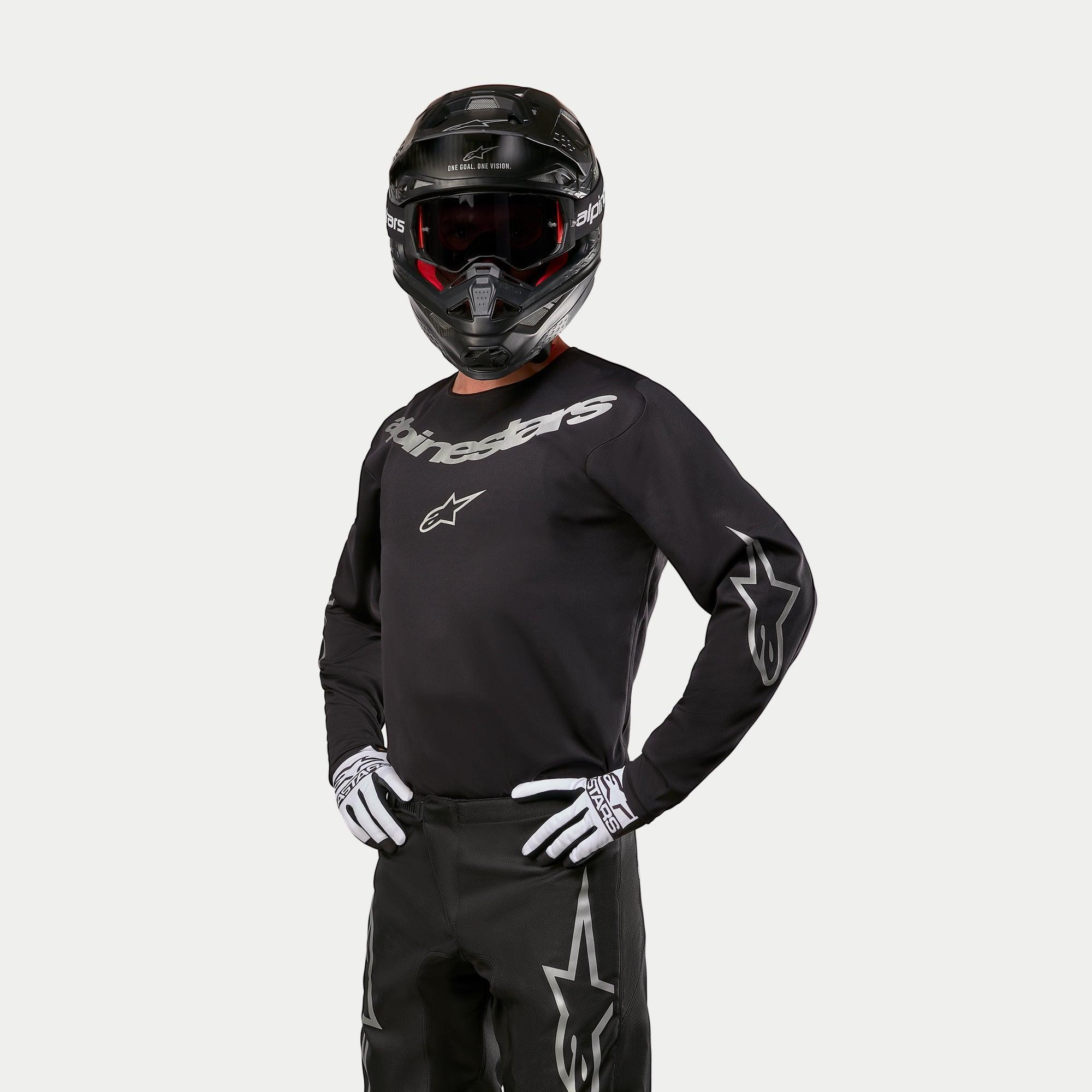 Pilote de motocross portant le maillot Alpinestars Fluid Graphite 2024 noir et argent, associé à un pantalon de motocross noir, un casque noir avec des lunettes teintées de rouge et des gants blancs et noirs, avec des logos argentés sur la poitrine et les manches.