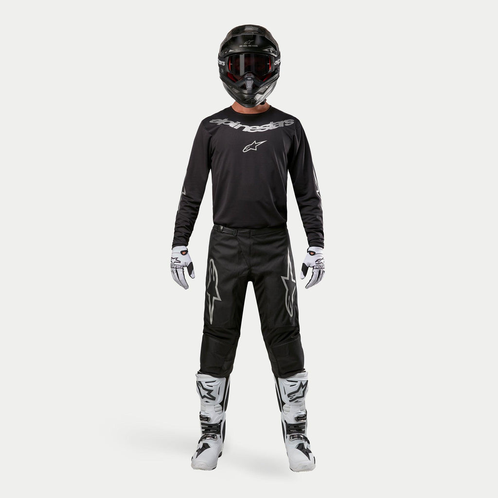 Pilote de motocross portant une tenue de course complète, maillot Alpinestars 2024 Fluid Graphite en Noir Argent, pantalon noir avec logos d'étoiles argentés, casque noir avec lunettes rouges, bottes blanches et noires, gants blancs et noirs, tenue de motocross coordonnée