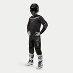 Pilote de motocross portant le maillot Alpinestars 2024 Fluid Graphite en noir et argent, pantalon noir assorti avec logos d'étoiles argentées, bottes blanches et noires, gants blancs et noirs, et casque noir avec lunettes rouges, équipement de course tout-terrain complet