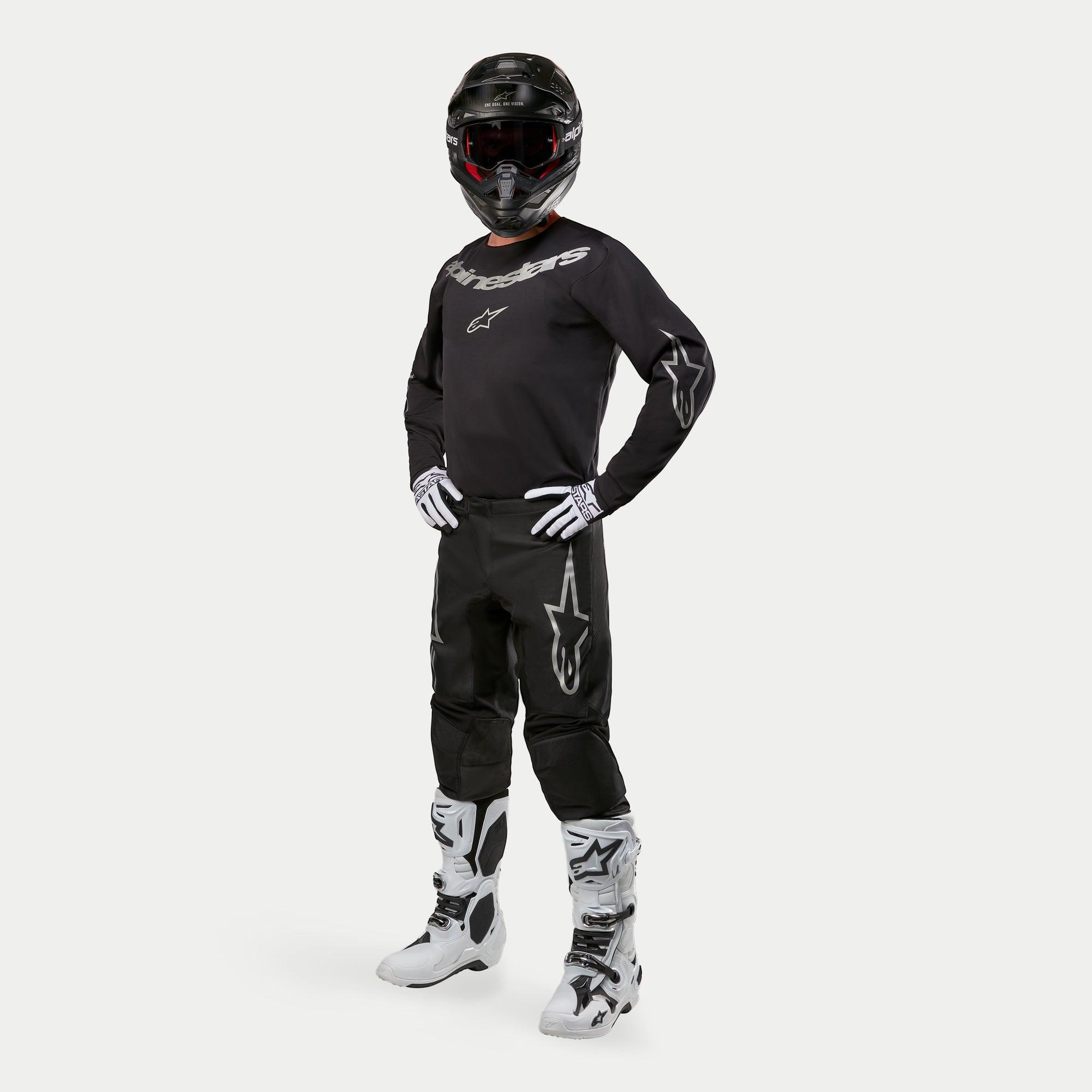 Pilote de motocross portant le maillot Alpinestars 2024 Fluid Graphite en noir et argent, pantalon noir assorti avec logos d'étoiles argentées, bottes blanches et noires, gants blancs et noirs, et casque noir avec lunettes rouges, équipement de course tout-terrain complet