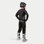 Pilote de motocross portant le maillot Alpinestars 2024 Fluid Graphite en Noir Argent, vu de dos, maillot à manches longues à coupe athlétique associé à un pantalon de motocross noir, un casque noir et des bottes blanches et noires, équipement professionnel complet pour la course tout-terrain.