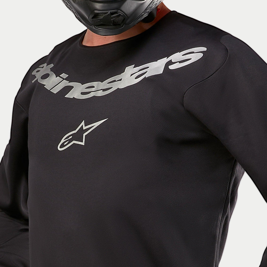 Maillot Alpinestars Fluid Graphite 2024, maillot de motocross, noir et argent, gros plan sur la poitrine avec marquage Alpinestars et logo étoile argentés, conception en poly-tissu respirant, porté par un pilote avec un casque noir