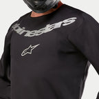 Maillot Alpinestars Fluid Graphite 2024, maillot de motocross, noir et argent, gros plan sur la poitrine avec marquage Alpinestars et logo étoile argentés, conception en poly-tissu respirant, porté par un pilote avec un casque noir