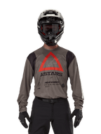 Motocross rider wearing Alpinestars Techdura Jersey in Falcon Brown, featuring a prominent red triangle and 'ASTARS' branding on the chest, paired with black pants, a black helmet with goggles, and black gloves, complete enduro racing outfit.
