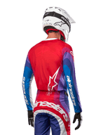 Pilote de motocross de dos portant le maillot Alpinestars 2024 Techstar Pneuma en Blue Mars Red White, avec un dos rouge orné d'un logo étoile blanc et des manches bleues et violettes avec marquage blanc, coordonné à un pantalon assorti et un casque blanc pour la course tout-terrain.