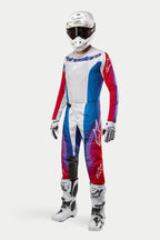 Pilote de motocross portant un équipement complet Alpinestars, pantalon Techstar Pneuma 2024 en bleu, rouge Mars et blanc, maillot à manches longues assorti, casque blanc avec lunettes sombres et bottes blanches et noires, tenue coordonnée pour la course tout-terrain