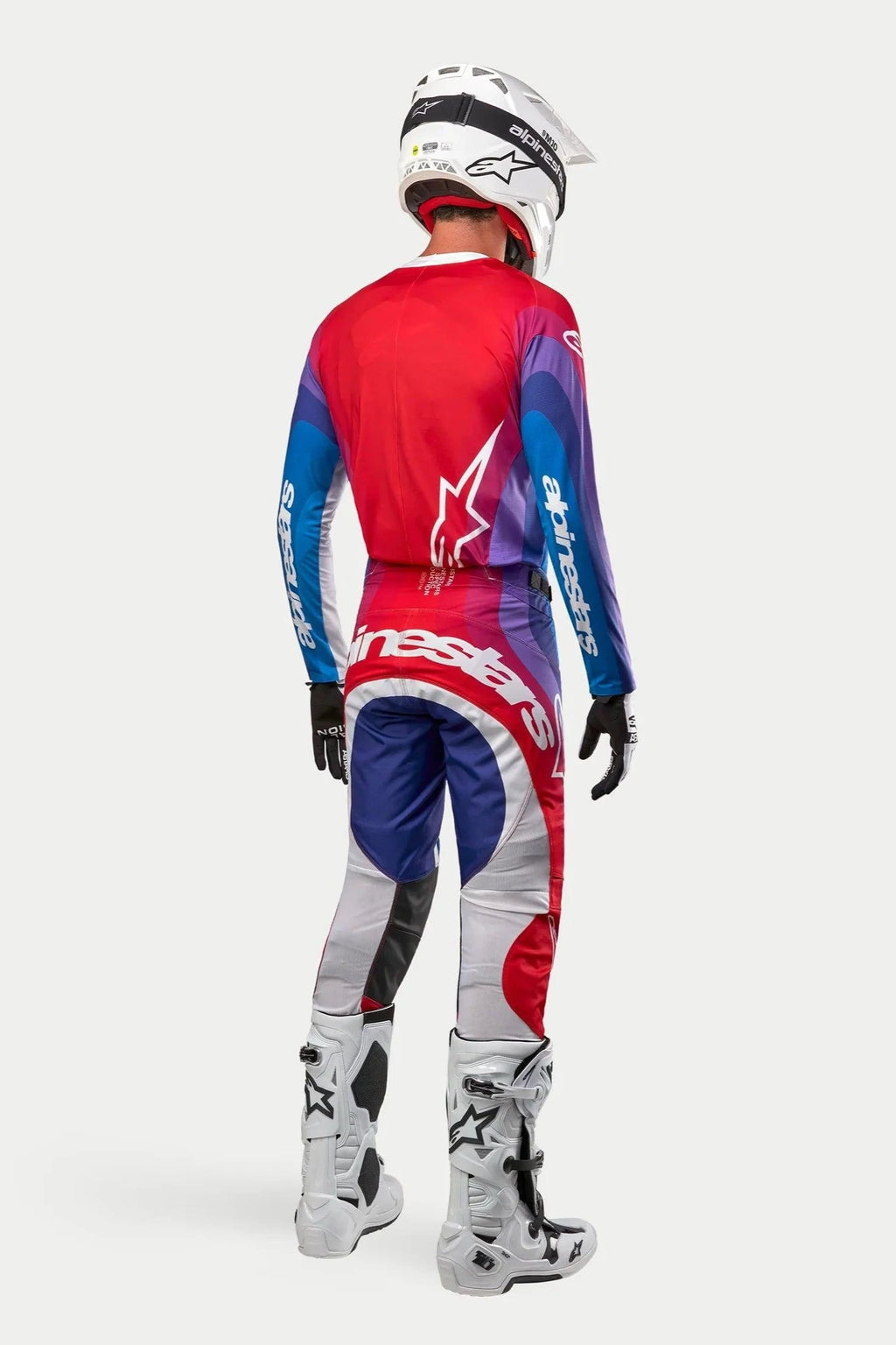 Pilote de motocross vu de dos portant le maillot et le pantalon Alpinestars Techstar Pneuma 2024, coloris Blu Mars Red White avec dégradés de rouge, bleu et violet, logos blancs, avec bottes et casque blancs, tenue de course tout-terrain coordonnée.