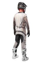 Pilote de motocross vu de dos portant le maillot Techstar Pneuma 2024 en Dark Sand Iron Dust Gray, pantalon de motocross assorti avec un logo imposant, casque noir et bottes de course blanches, équipement tout-terrain professionnel coordonné.