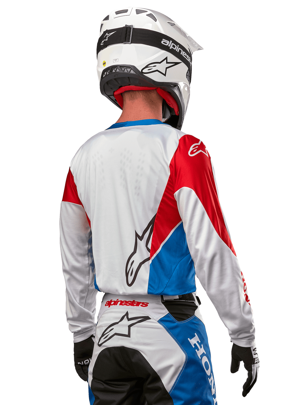 Pilote de motocross portant le maillot Alpinestars Honda Racer Iconic, vue arrière, blanc avec empiècements bleu vif et rouge vif, logo Alpinestars proéminent au dos, associé à un casque de motocross blanc et un pantalon de course assorti.
