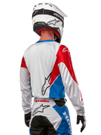 Pilote de motocross portant le maillot Alpinestars Honda Racer Iconic, vue arrière, blanc avec empiècements bleu vif et rouge vif, logo Alpinestars proéminent au dos, associé à un casque de motocross blanc et un pantalon de course assorti.