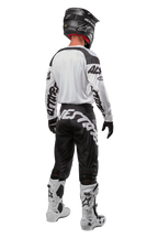 Maillot Alpinestars Racer Hana 2024, vue arrière d'un pilote de motocross, blanc et noir, maillot blanc avec marquage graphique noir sur les manches, associé à un pantalon noir avec lettrage Alpinestars blanc sur l'assise, casque noir et bottes blanches et noires, équipement de course tout-terrain coordonné