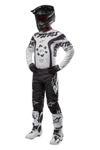 Pilote de motocross portant une tenue complète avec le maillot Alpinestars Racer Hana 2024 en blanc et noir, un pantalon de motocross noir avec des logos blancs, un casque noir, des bottes de course blanches et noires et des gants noirs, tenue de course tout-terrain professionnelle