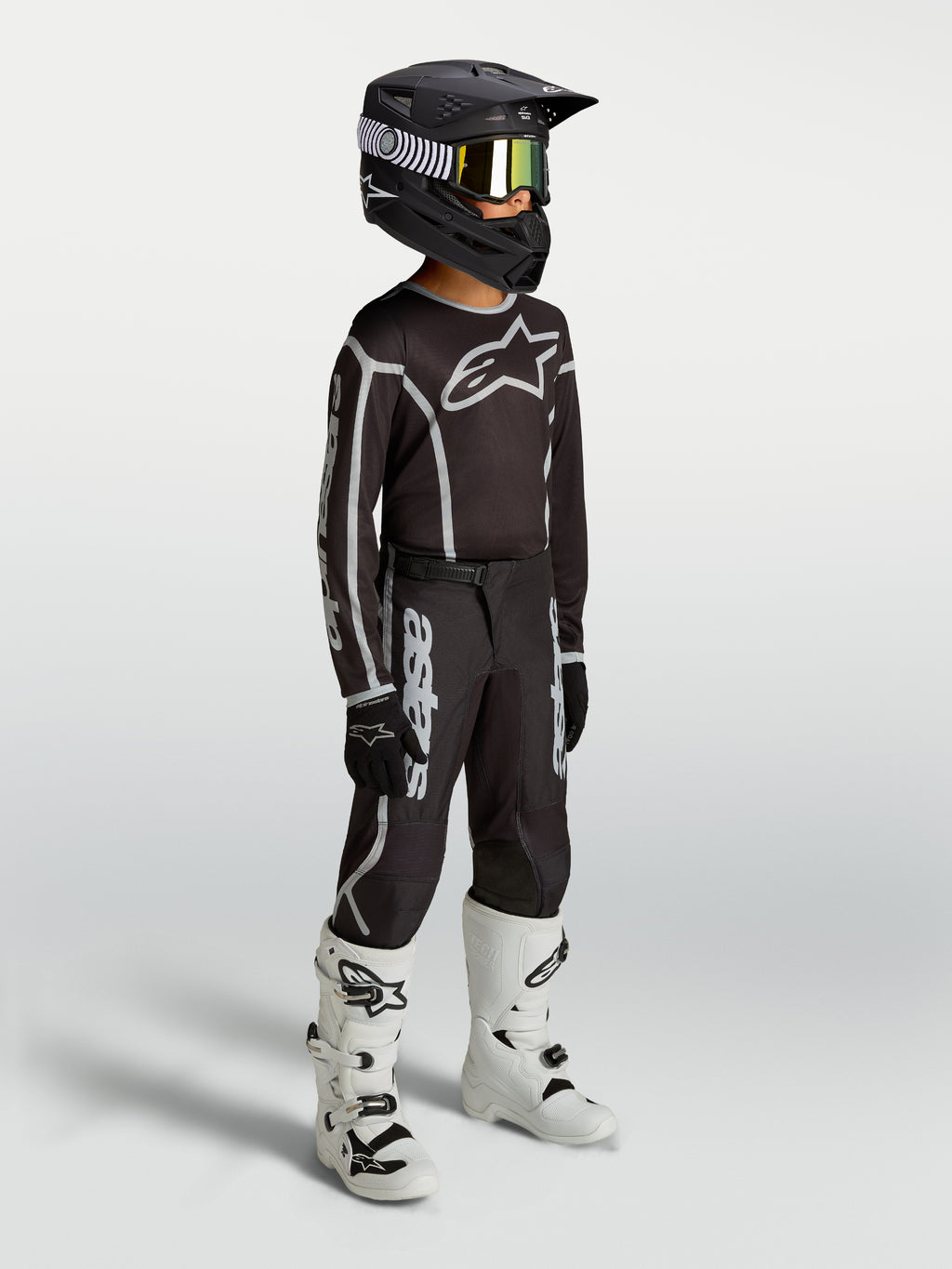 Alpinestars maillot Fluid Apex V2 Junior et ensemble de motocross pour enfant, noirs et argentés, textile respirant, porté par un jeune pilote avec des bottes blanches.