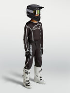 Alpinestars maillot Fluid Apex V2 Junior et ensemble de motocross pour enfant, noirs et argentés, textile respirant, porté par un jeune pilote avec des bottes blanches.