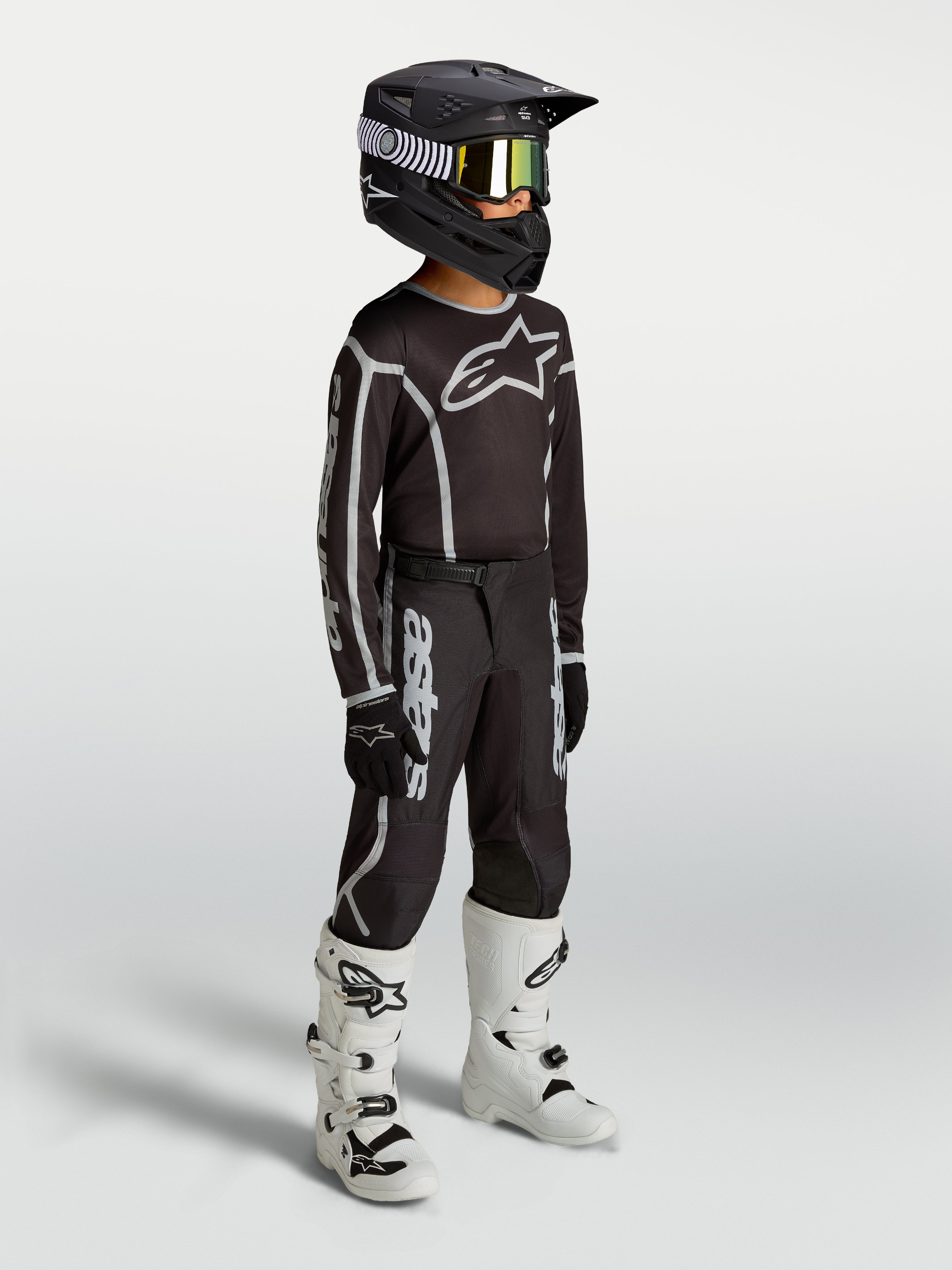 Alpinestars maillot Fluid Apex V2 Junior et ensemble de motocross pour enfant, noirs et argentés, textile respirant, porté par un jeune pilote avec des bottes blanches.