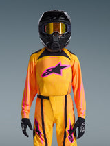 Jeune pilote de motocross portant le Maillot Supertech Nomur Junior 2026, dégradé orange et jaune avec logo Astars rose vif, pantalon assorti avec accents noirs et roses, casque noir avec lunettes à verres dorés et gants noirs, ensemble d'équipement de course tout-terrain professionnel