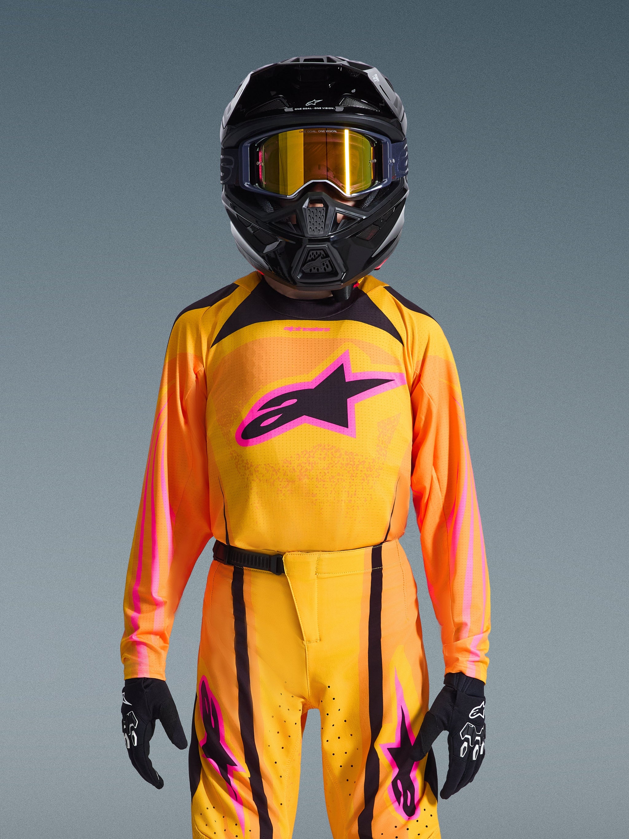 Jeune pilote de motocross portant le Maillot Supertech Nomur Junior 2026, dégradé orange et jaune avec logo Astars rose vif, pantalon assorti avec accents noirs et roses, casque noir avec lunettes à verres dorés et gants noirs, ensemble d'équipement de course tout-terrain professionnel
