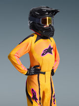 Jeune pilote de motocross portant le maillot Alpinestars Junior Supertech Nomur 2026, orange, jaune et rose, pantalon de course assorti, casque noir avec lunettes et gants noirs, coupe athlétique, équipement tout-terrain haute performance