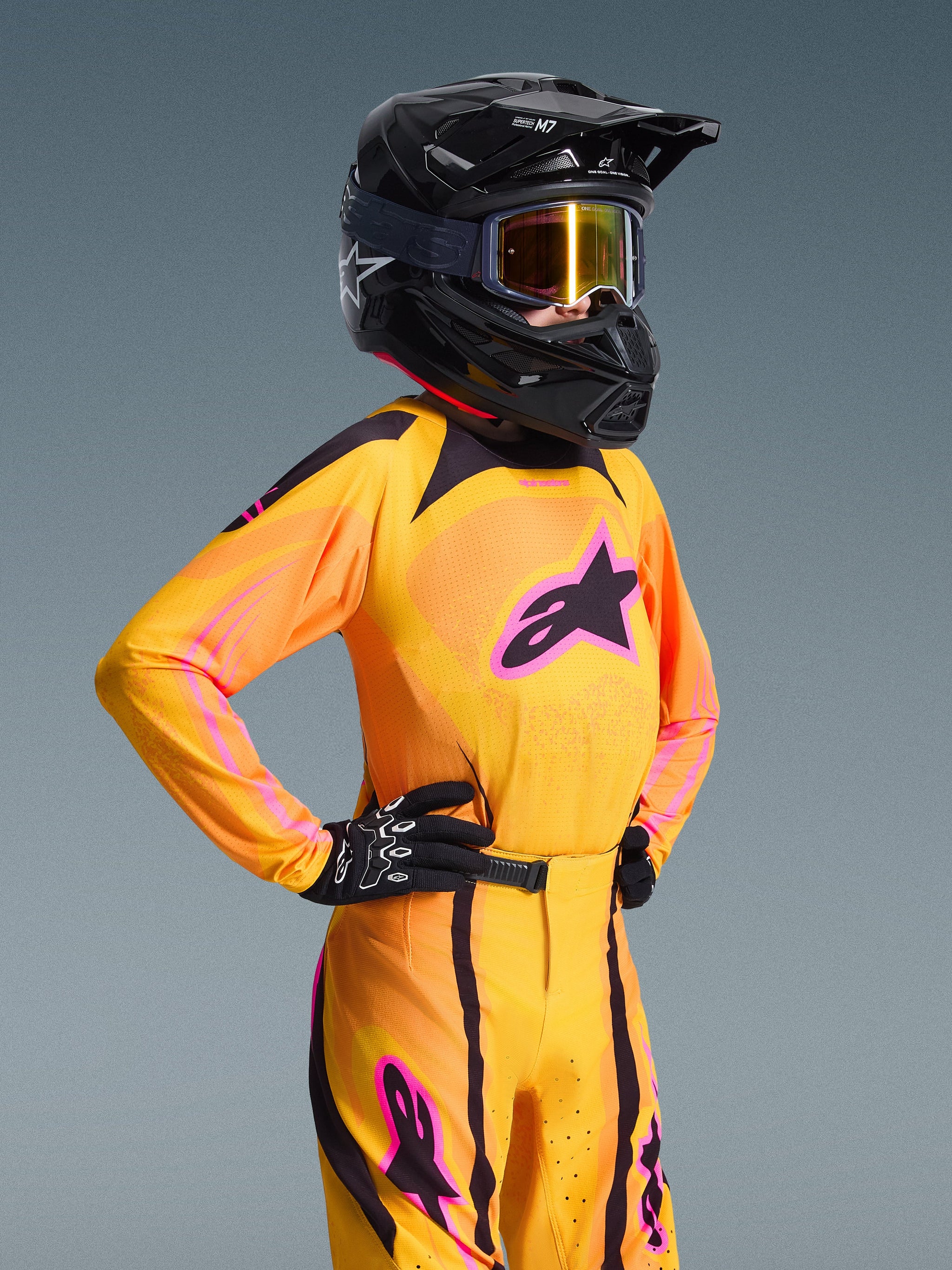 Jeune pilote de motocross portant le maillot Alpinestars Junior Supertech Nomur 2026, orange, jaune et rose, pantalon de course assorti, casque noir avec lunettes et gants noirs, coupe athlétique, équipement tout-terrain haute performance