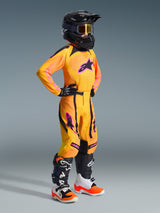 Jeune pilote de motocross portant une tenue de course complète, Maillot Junior Supertech Nomur orange avec des accents jaunes et roses, pantalon de motocross assorti, casque noir avec lunettes, et bottes noires et blanches, tenue de niveau pro pour la course tout-terrain