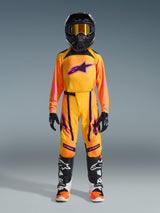 Jeune pilote de motocross portant un équipement de course complet, Maillot Alpinestars Junior Supertech Nomur 2026 en orange, jaune et rose, pantalon de motocross assorti, casque noir avec lunettes à effet miroir orange, gants noirs et bottes de course techniques, tenue professionnelle coordonnée pour la course tout-terrain