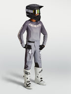 Ensemble Alpinestars Fluid Amber Junior, maillot et pantalon de motocross gris, bottes de course blanches et casque noir.