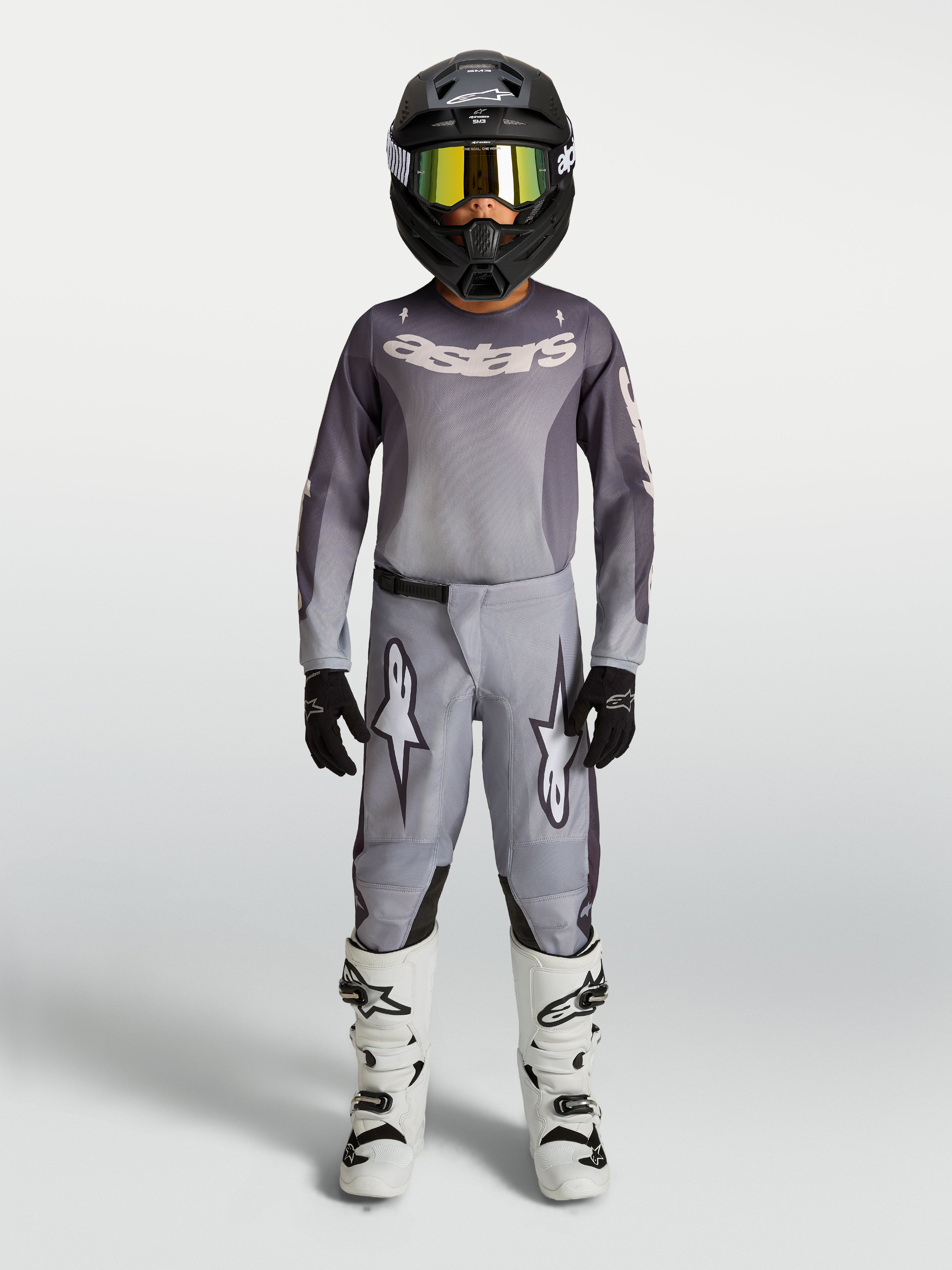 Alpinestars maillot et ensemble Fluid Amber junior 2027, tenue de motocross grise et sable, casque noir et bottes blanches.