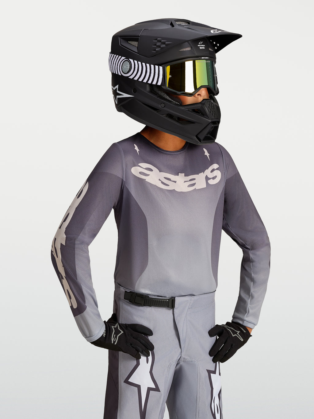 Maillot Fluid Amber Junior 2027 d'Alpinestars, maillot de motocross gris et sable pour enfant, porté avec un casque de moto noir et des gants.