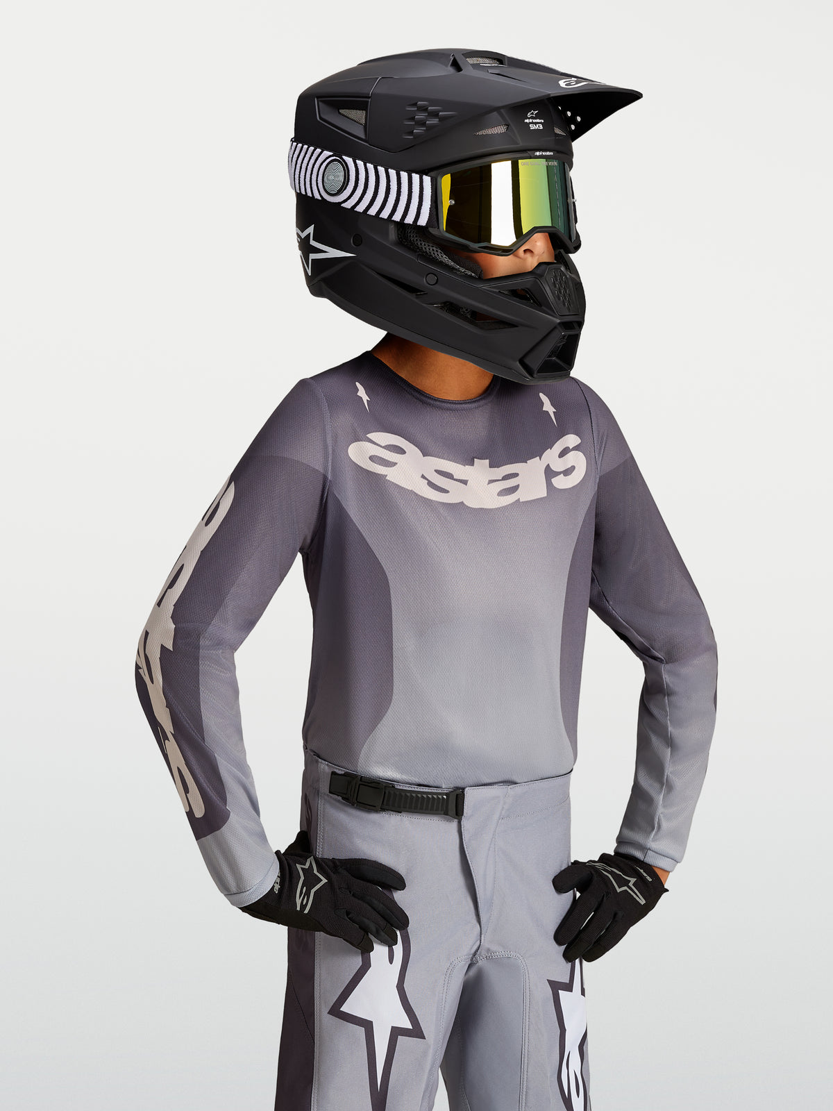 Maillot Fluid Amber Junior 2027 d'Alpinestars, maillot de motocross gris et sable pour enfant, porté avec un casque de moto noir et des gants.