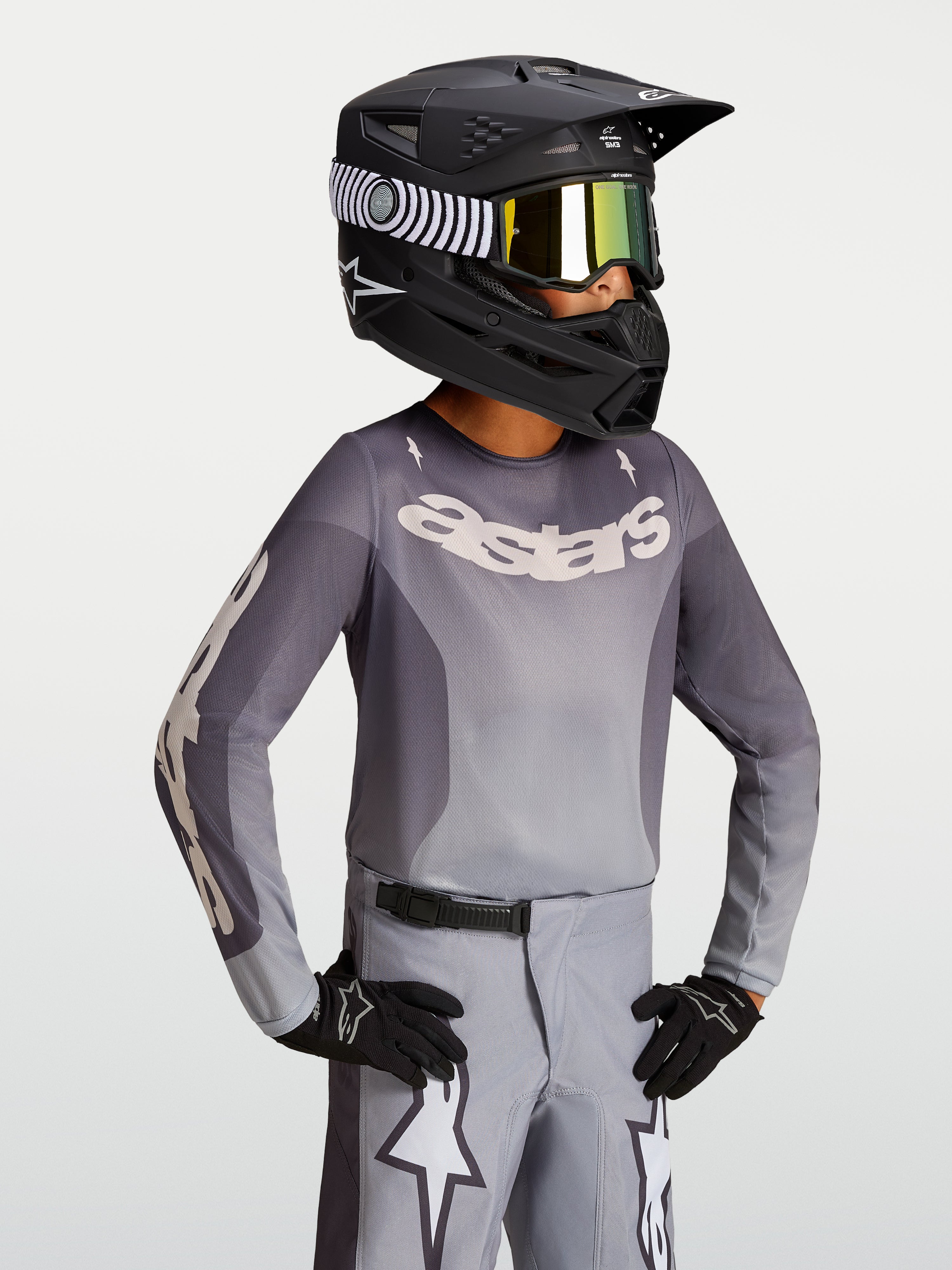 Maillot Fluid Amber Junior 2027 d'Alpinestars, maillot de motocross gris et sable pour enfant, porté avec un casque de moto noir et des gants.